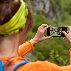 Backwards shot of woman tourist makes photo of river in ravine on smartphone device for posting in social networks, makes video for chanel, carries backpack, amazing nature view. Place for inscription